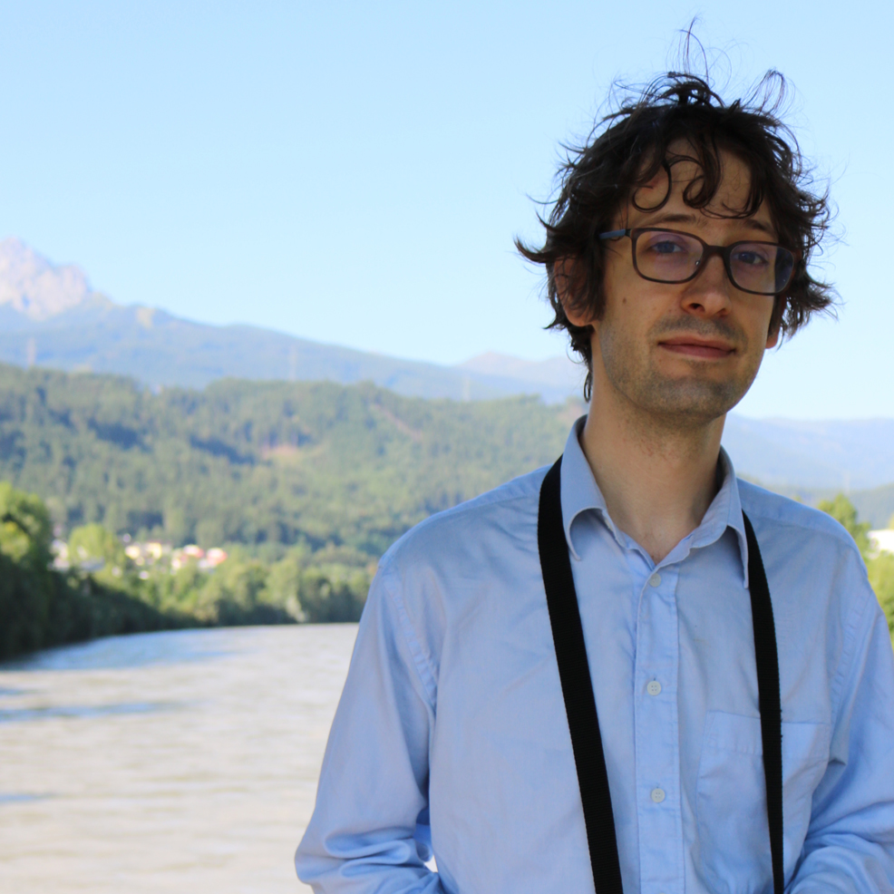 Me in 2017, unshaven, messy hair, square glasses, blue shirt. Background is sunny, has a gray river, forest, part of a mountain, blue sky